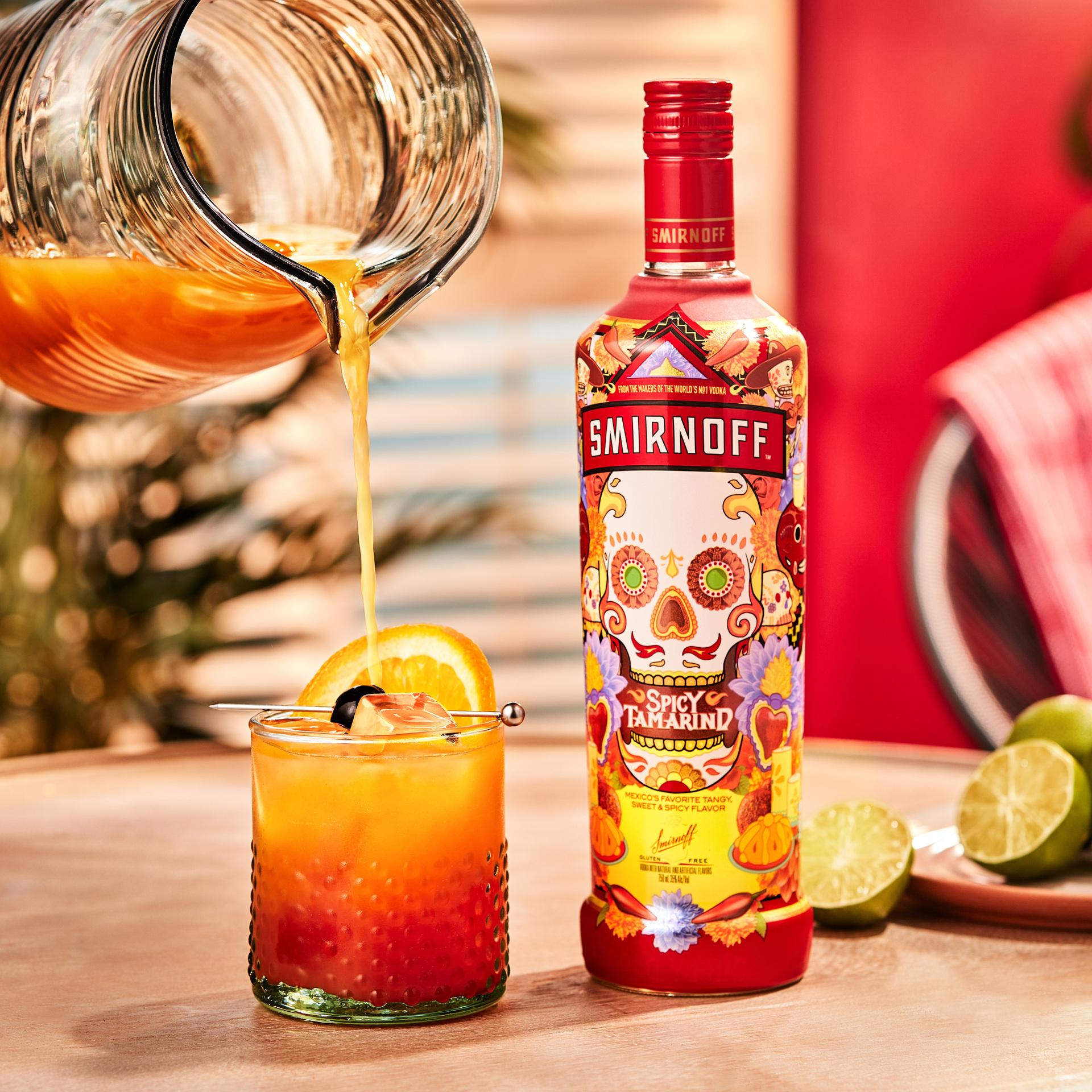 Smirnoff Spicy Tamarind vodka bottle alongside a pitcher pouring a Spicy Sunrise cocktail with an orange slice and maraschino cherry for garnish. 
