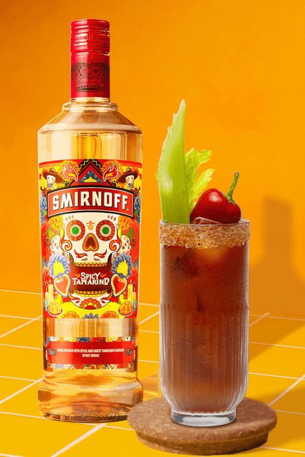 Smirnoff Spicy Tamarind vodka bottle beside a Bloody Mary cocktail with a chili powder rim, garnished with a celery stalk and a red chili pepper, set against a vibrant yellow background with white grid lines.