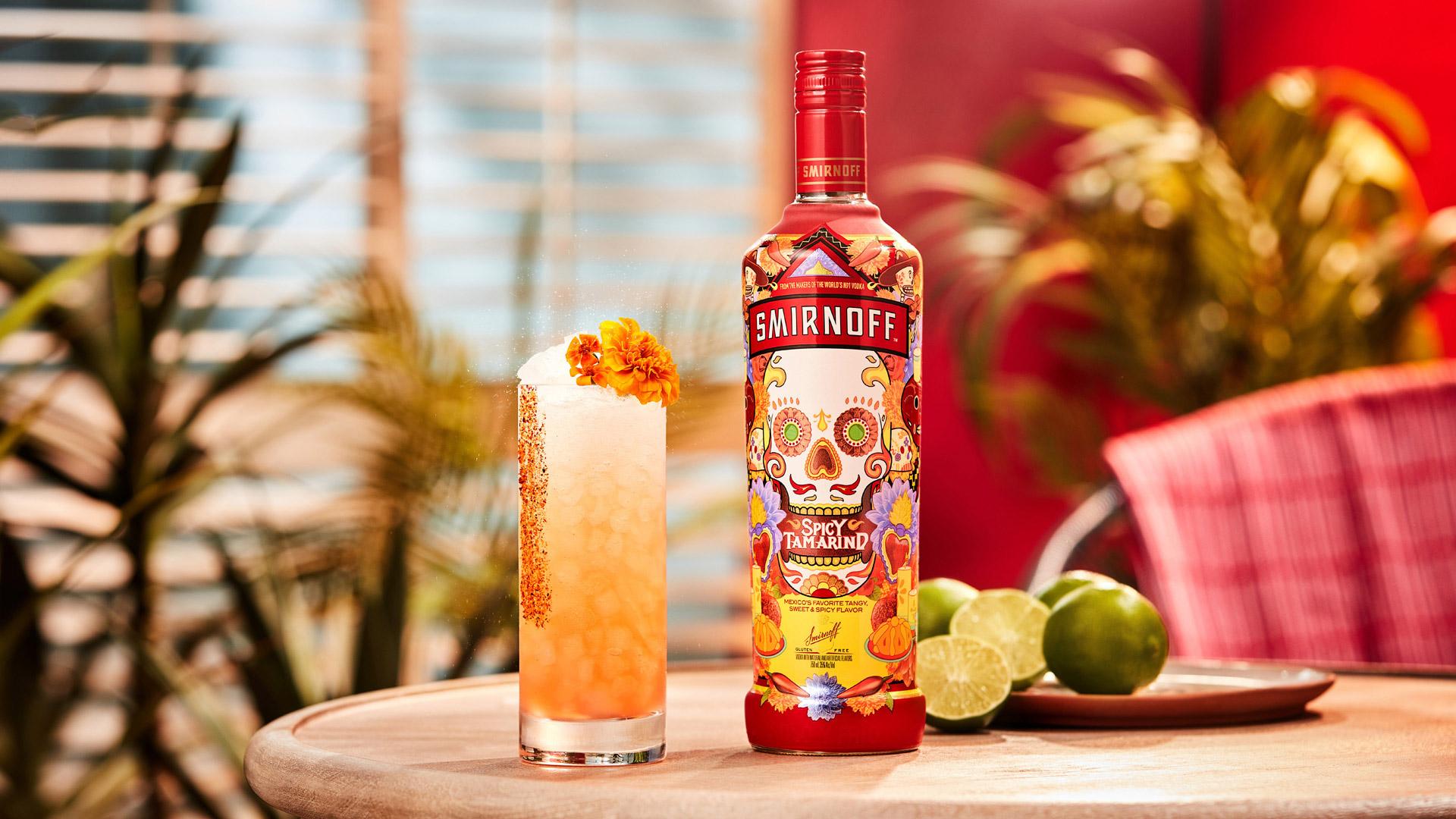 Smirnoff Spicy Tamarind vodka bottle alongside an orange colored Sweet 'N' Spicy cocktail with spicy seasoning and marigold flower garnish. 