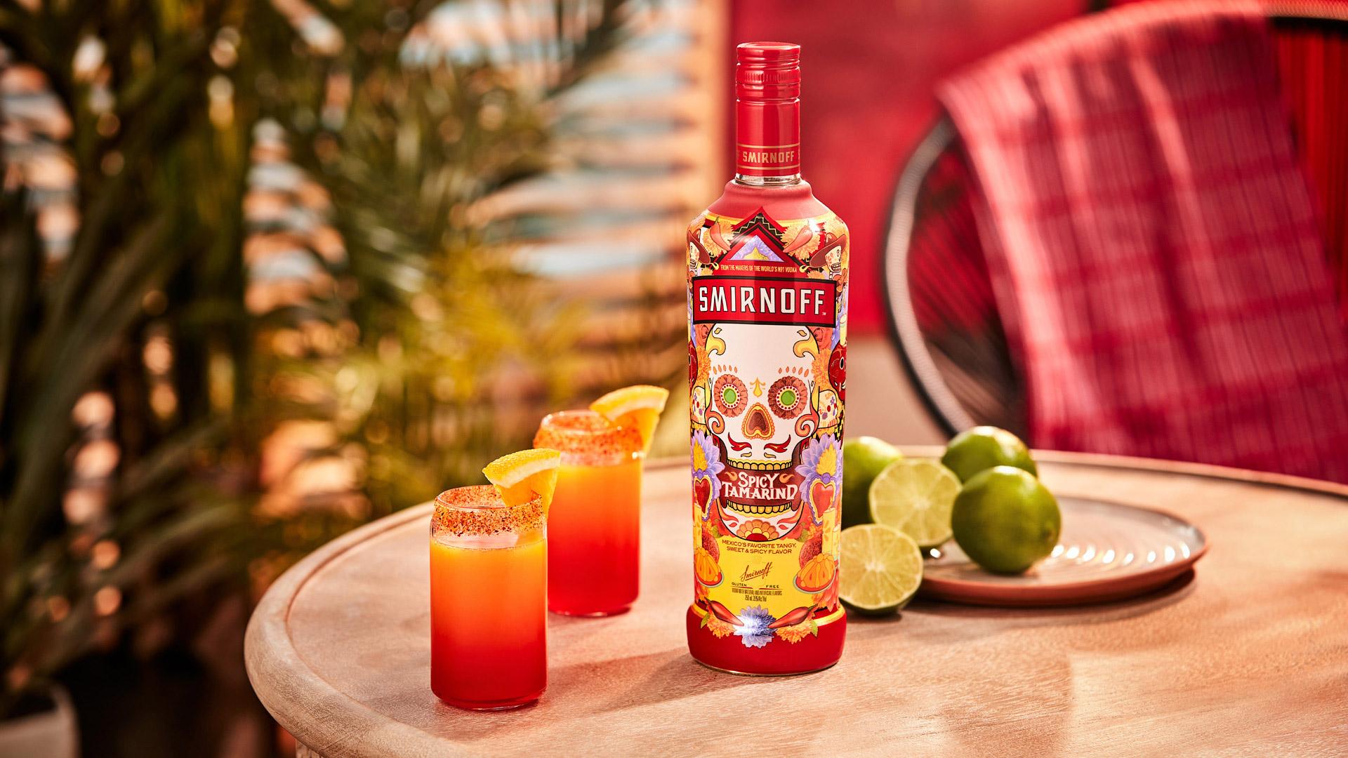 Smirnoff Spicy Tamarind vodka bottle alongside two orange and red colored Real Macoy Chamoy cocktails  with a spicy seasoning rim and orange wedge garnish. 