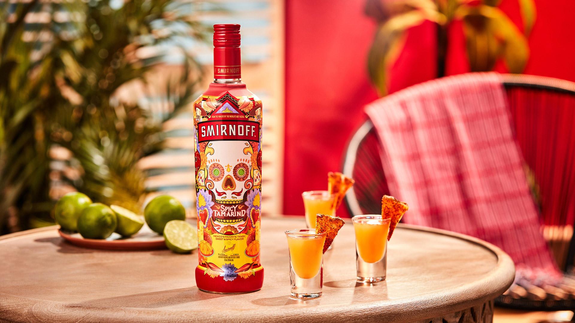 Smirnoff Spicy Tamarind vodka bottle alongside three orange colored Pineapple Drop Shots with spicy seasoning pineapple wedges for garnish. 